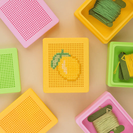 Flat lay of colorful cross stitch boxes with stitchable grid lids, featuring a finished lemon cross stitch design on one box and blank embroidery box lids arranged around it.