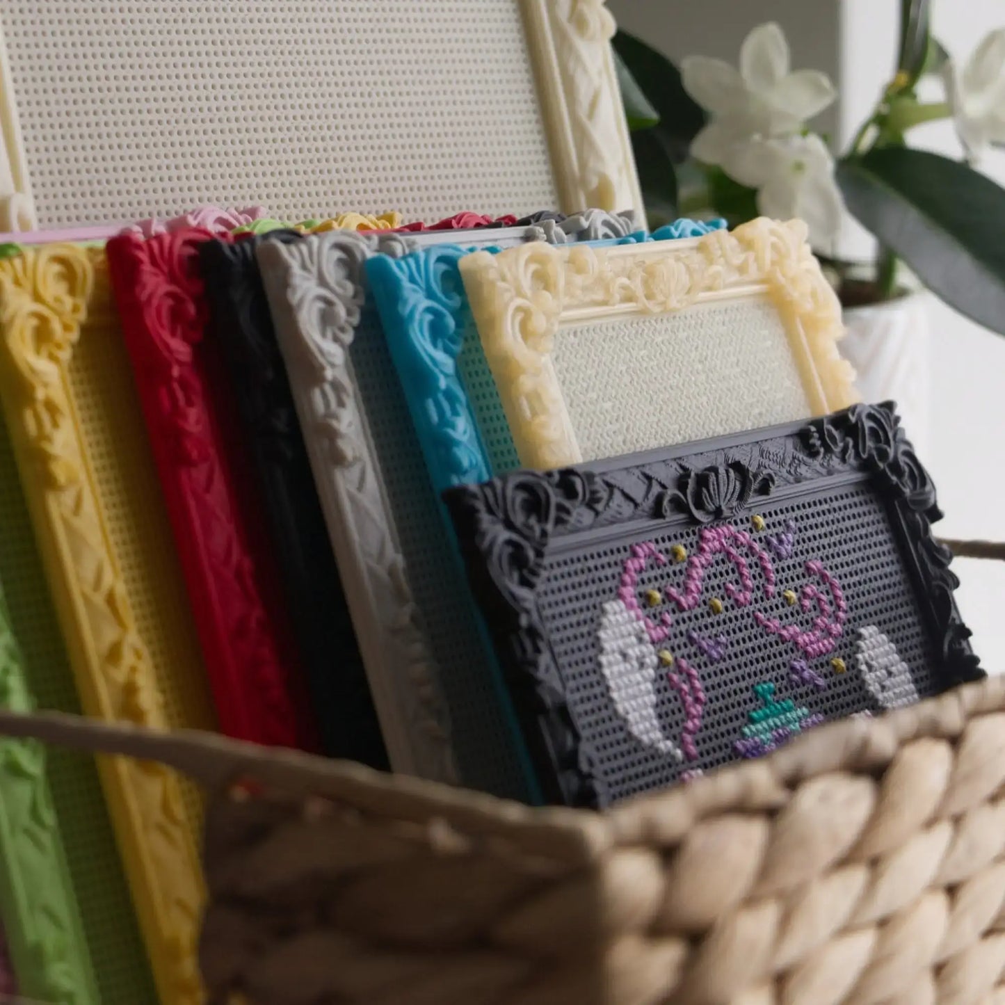 Baroque rectangle cross stitch frames in yellow, red, black, grey, blue and cream stacked in a woven basket, with a finished ghost teapot design in front.