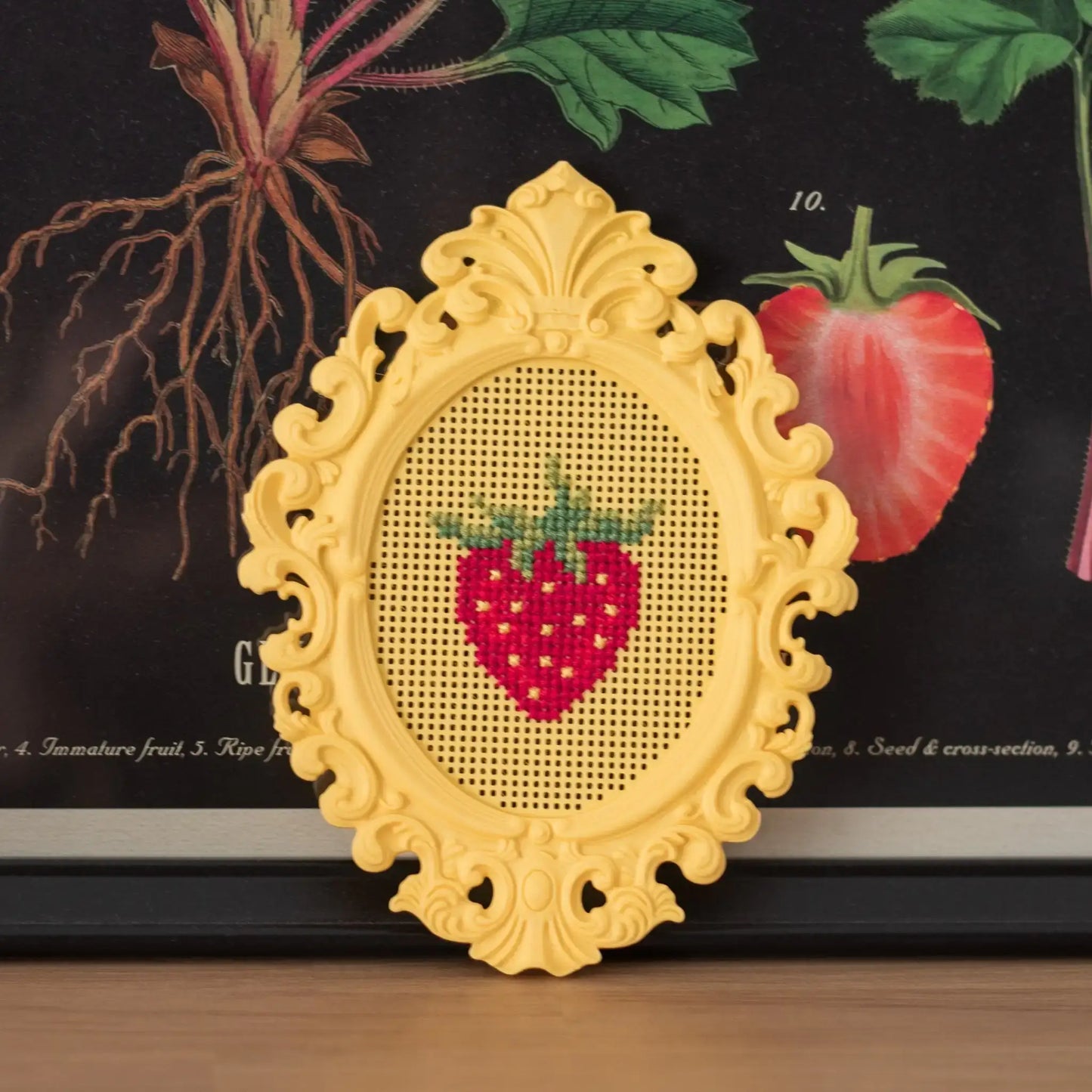 Stitchable cross stitch frame with a finished strawberry design, shown as a decorative display against a botanical illustration background.
