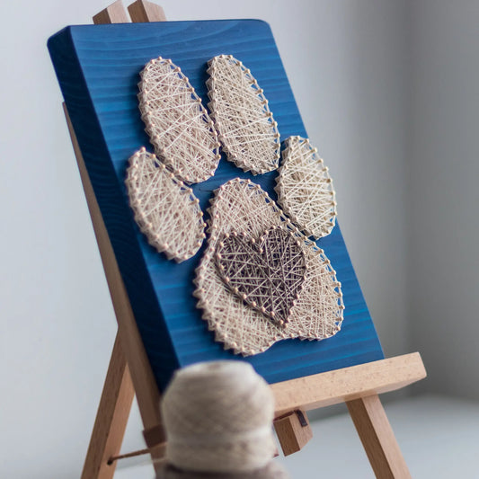 charming dog paw print string art sign made with white and dark brown string on a blue wooden background, featuring a heart shape in the center of the paw print.