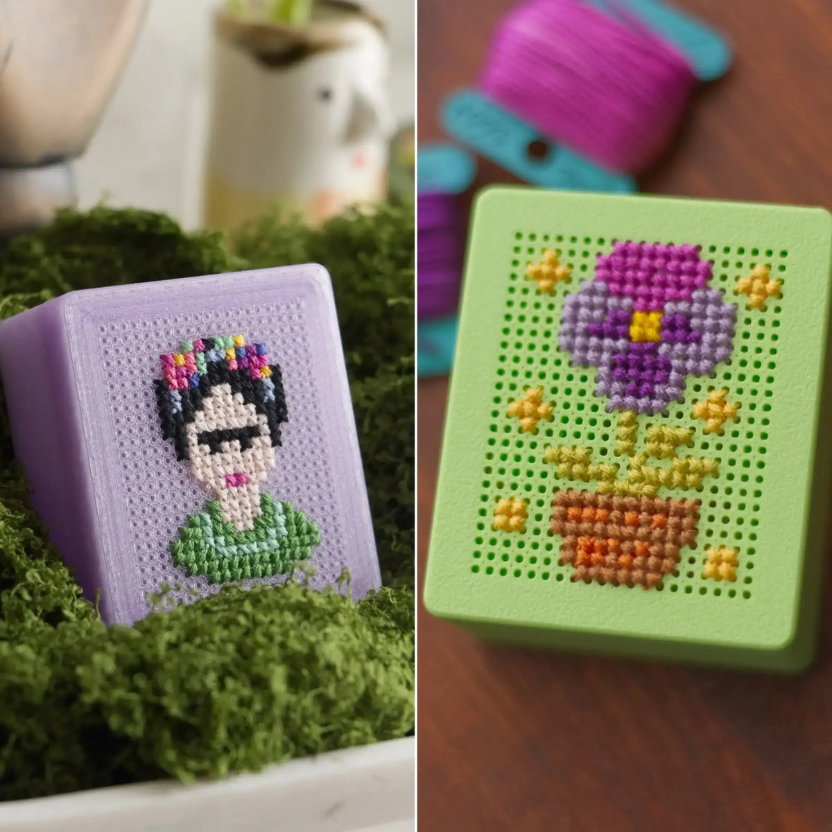 Two finished 3D-printed cross stitch boxes side by side: a lavender box with a Frida-inspired portrait design nestled in green moss, and a light green box with a purple flower and pot design on a wood surface.