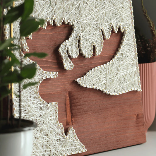 Finished moose silhouette string art made with ivory thread and nails on a stained wooden base, styled with plants to show the DIY string art kit as natural home decor.