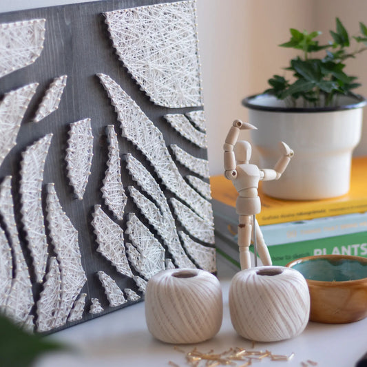 ree roots string art displayed alongside crafting materials like string spools and a wooden mannequin figure. The minimalist design complements the workspace decor.