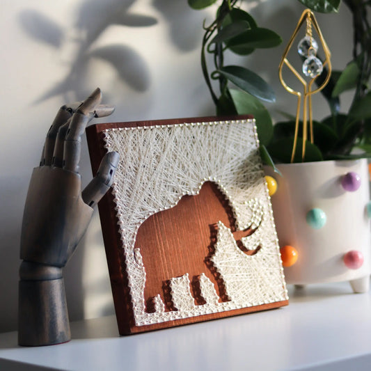 Artfully displayed DIY woolly mammoth string art kit on a wooden base, paired with a decorative plant and a wooden hand, showcasing a cozy and artistic setup.