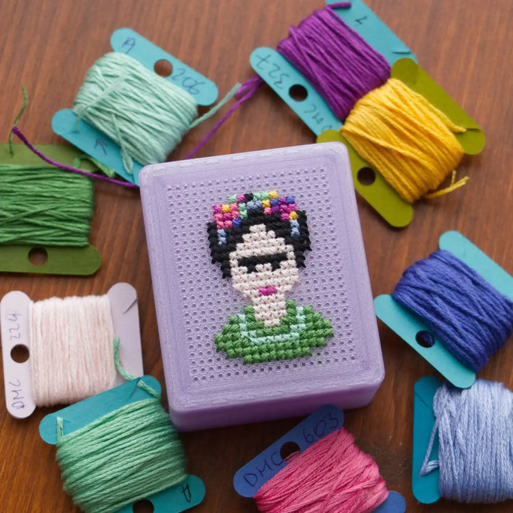 Light purple 3D-printed cross stitch box with a Frida-inspired portrait design stitched into the perforated lid, surrounded by bobbins of embroidery floss in green, yellow, purple, and blue on a wood surface.