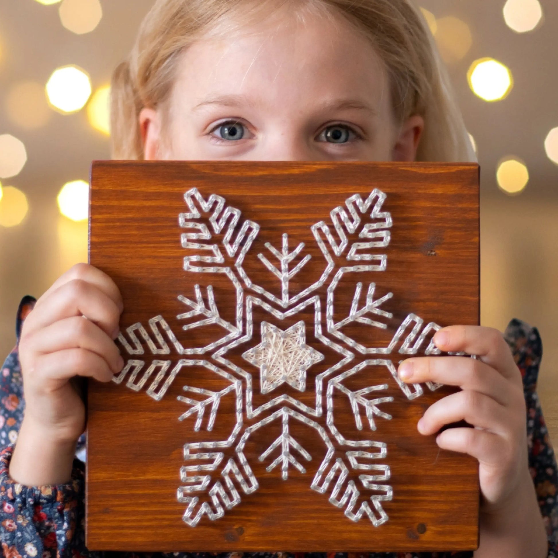 A young girl holding a wooden board displaying a snowflake string art design with ivory-colored threads, smiling with a blurred background.