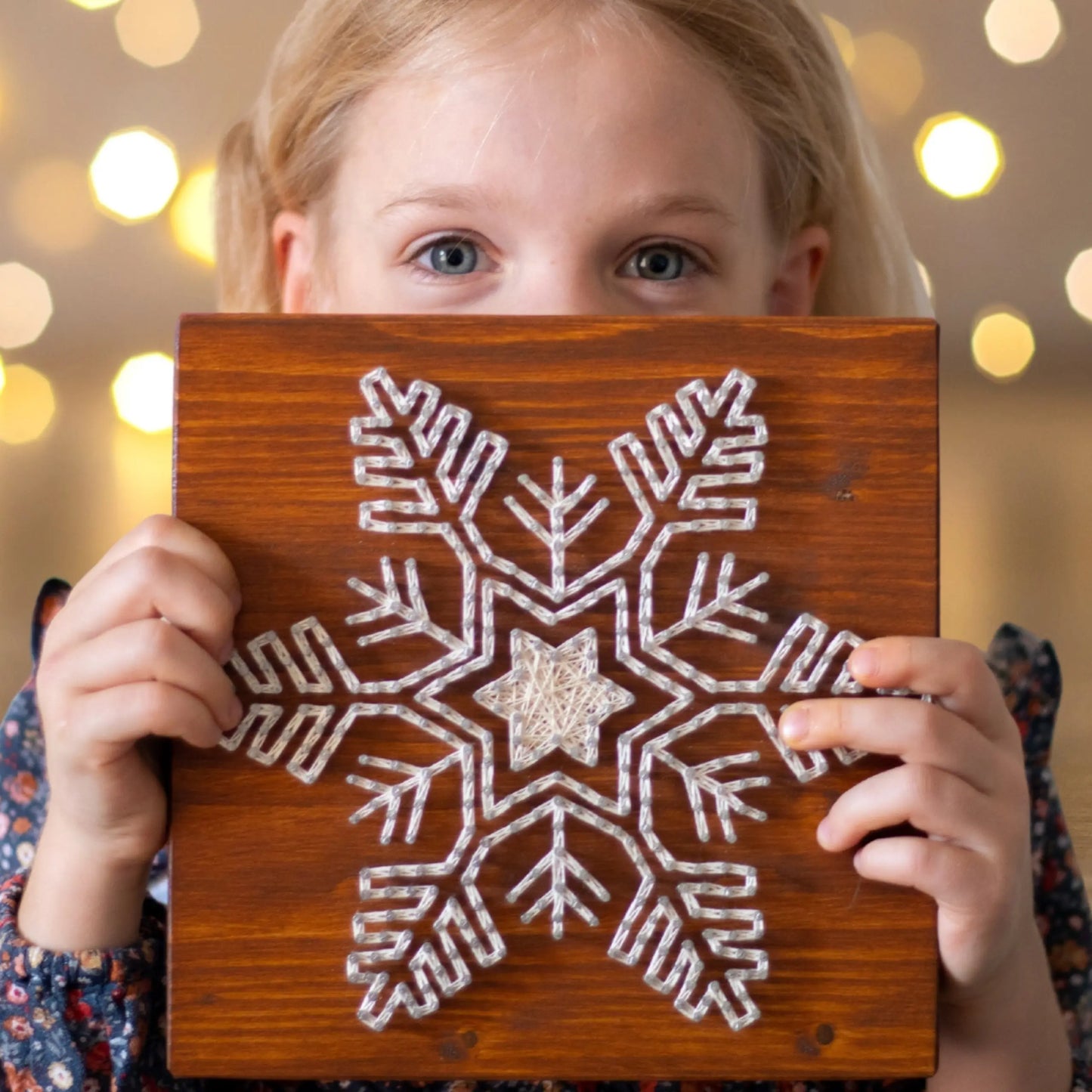 A child holding a finished snowflake string art sign, crafted with GoodStrings' snowflake string art pattern, showcasing the intricate white thread design on a wooden background.