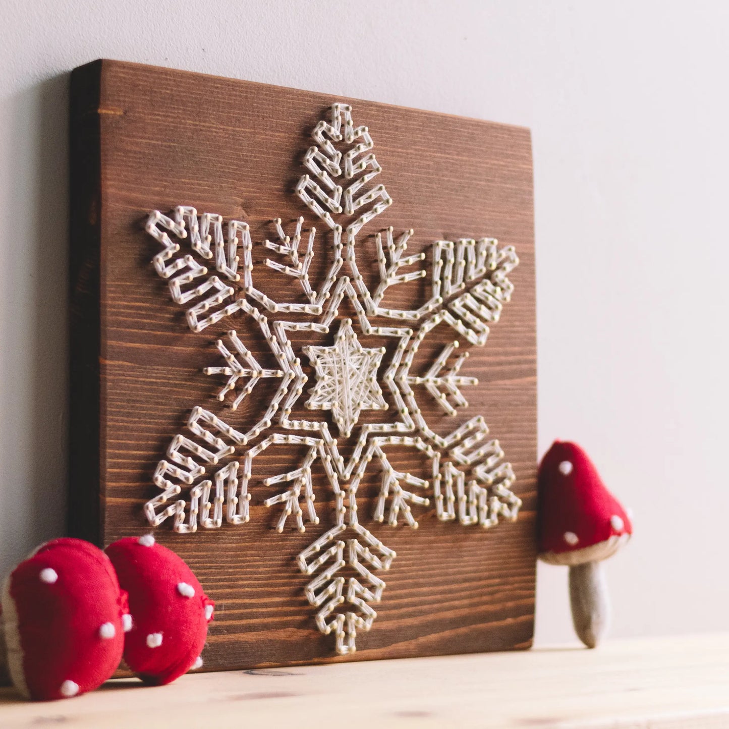 Side view of a finished snowflake string art piece on a wooden board, with red mushroom decorations on a wooden surface for a festive look.