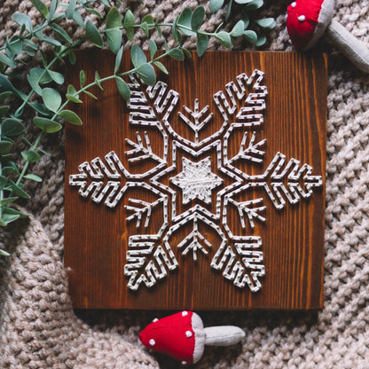 A snowflake string art piece made with ivory-colored threads on a wooden board, surrounded by greenery and fabric, with mushroom decorations.