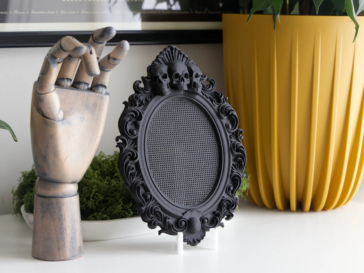 Black gothic cross stitch frame with three skulls beside a wooden hand and a yellow planter