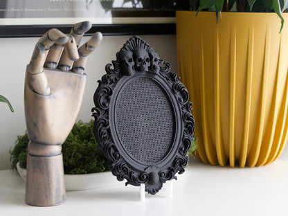 Black gothic cross stitch frame with three skulls beside a wooden hand and a yellow planter