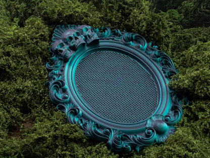 Teal three skulls cross stitch frame lying flat on green moss, showing the full oval grid