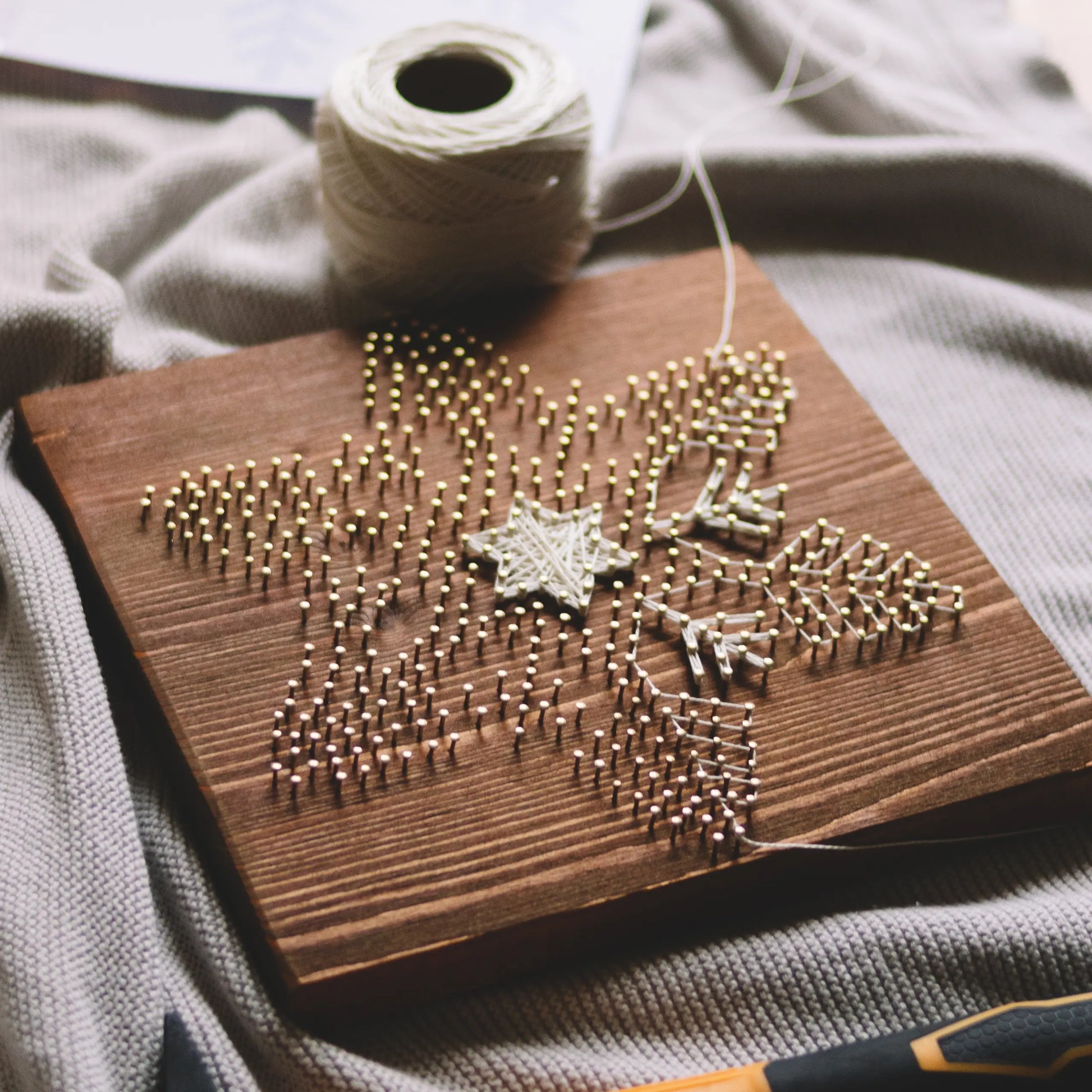 A partially completed snowflake string art piece on a wooden board, with a spool of ivory thread and a tool on a soft fabric background.