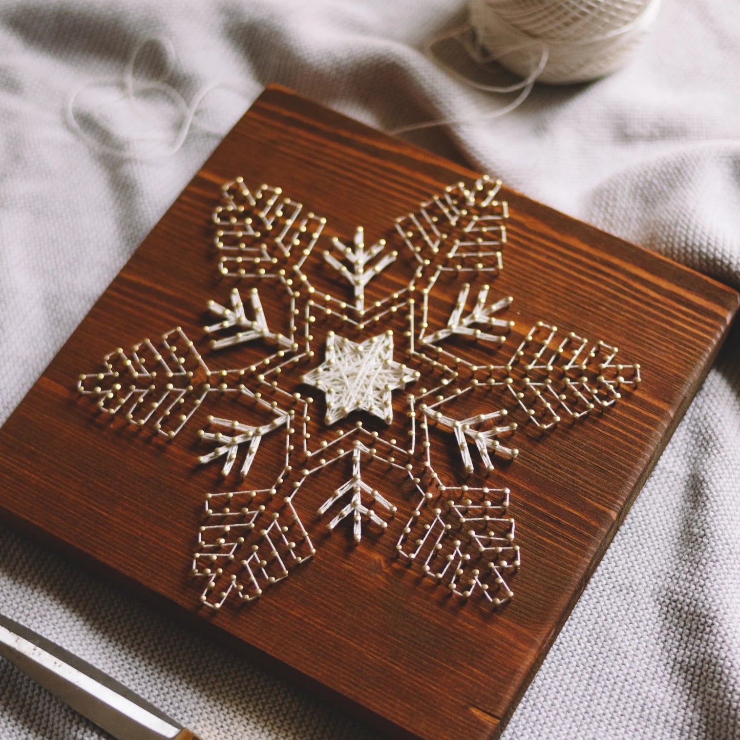 A nearly finished snowflake string art design on a wooden board, with threads in progress and a spool of thread on a soft fabric surface.