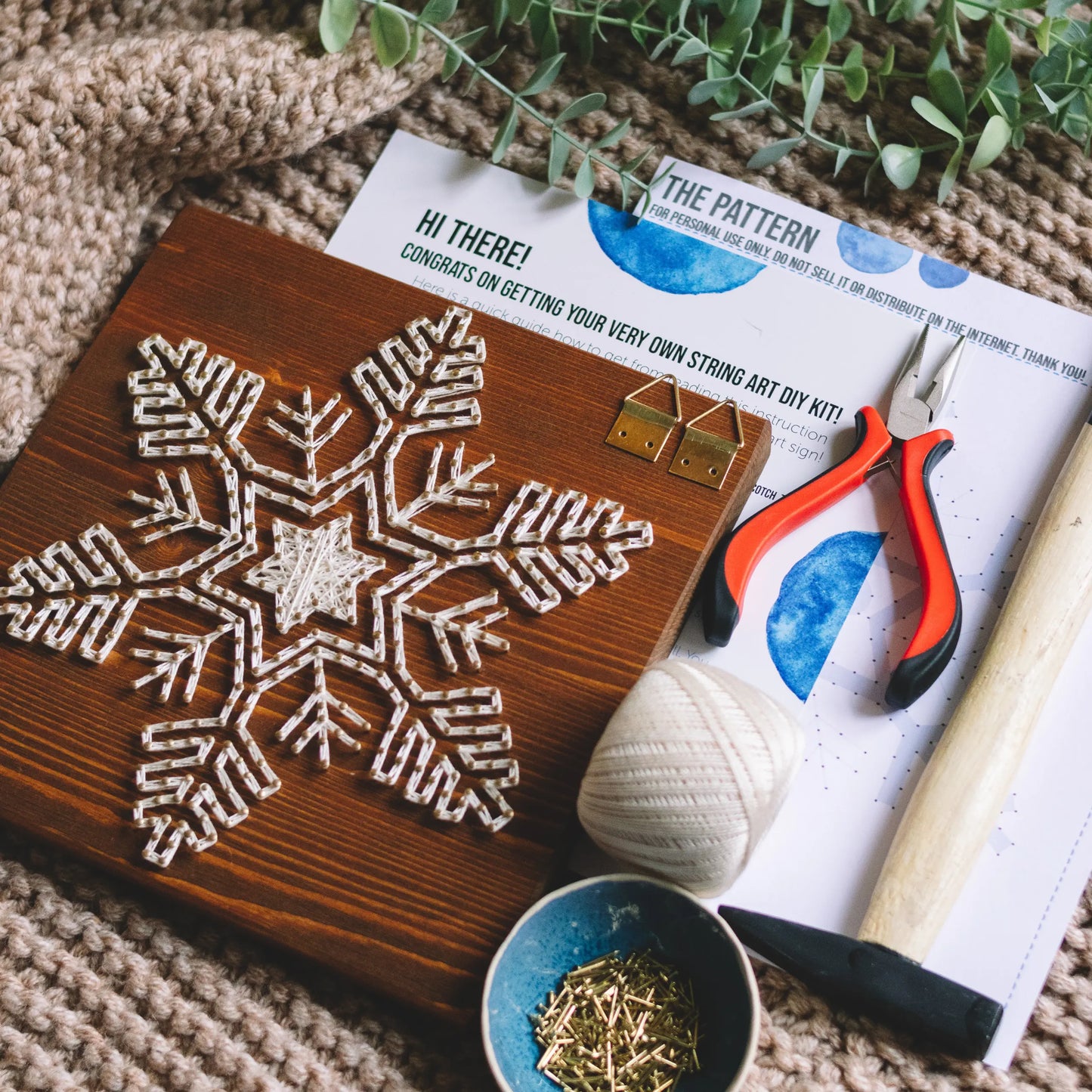 A completed snowflake string art kit with tools and instructions displayed on a cozy knitted blanket, surrounded by greenery and crafting tools.