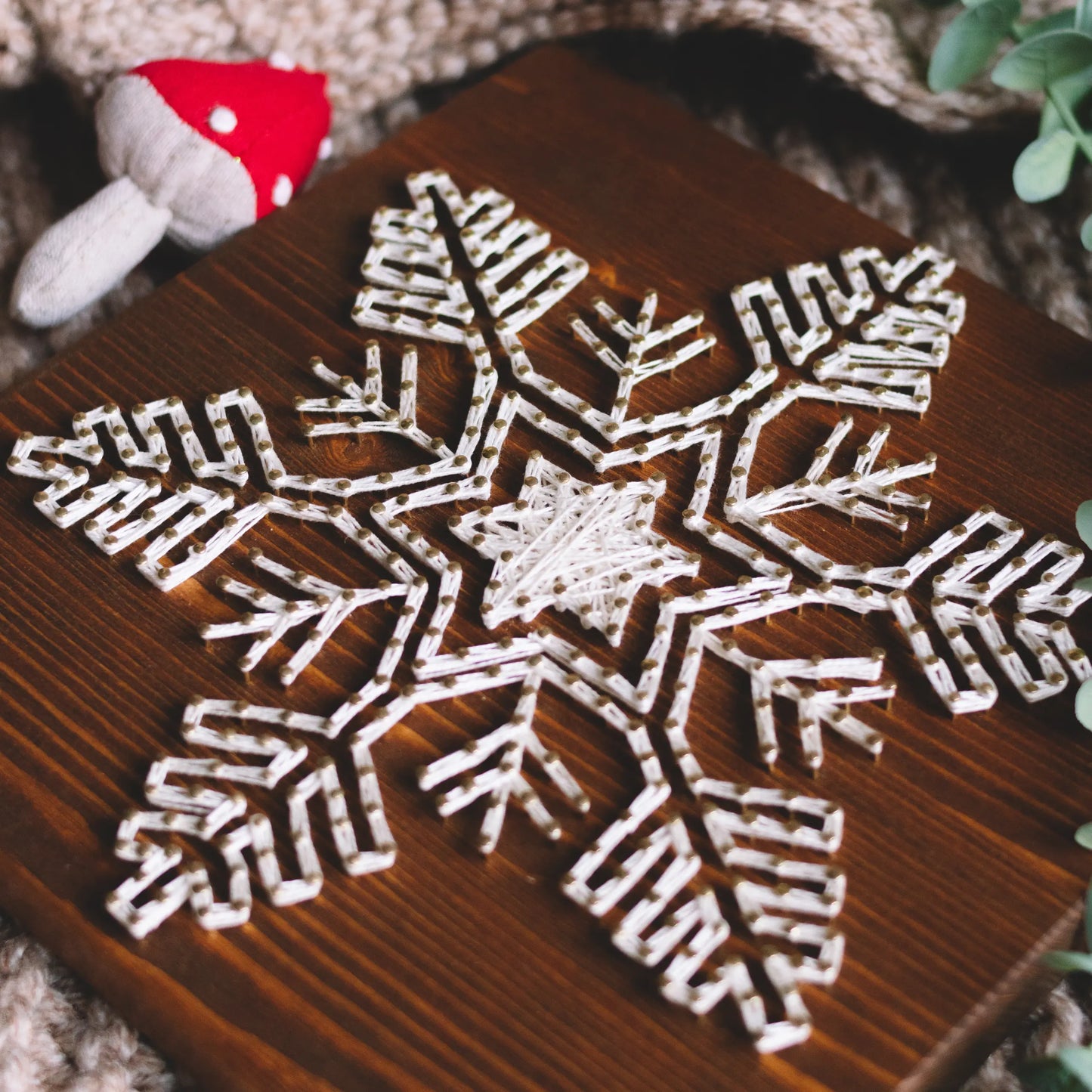 A completed snowflake string art piece made with ivory threads on a dark wood base, accompanied by red mushroom decorations on a soft surface.