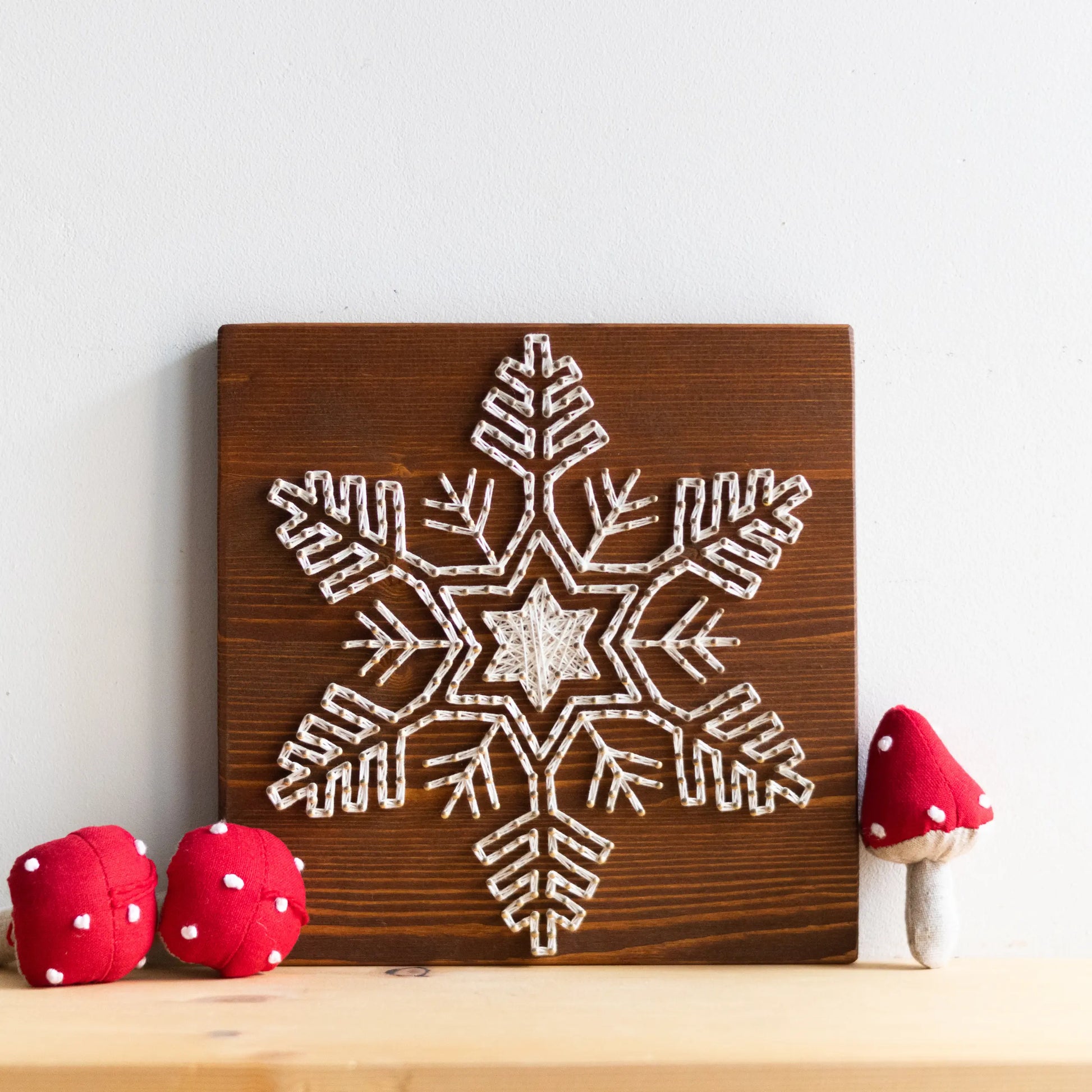 A finished snowflake string art piece displayed on a shelf with small red mushroom decorations, creating a cozy winter-themed decoration.