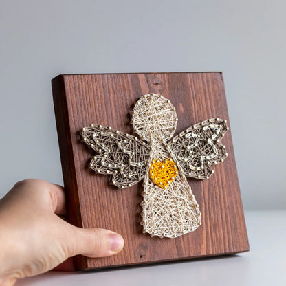 Wooden plaque with a string art angel holding a heart, held by a hand against a gray background