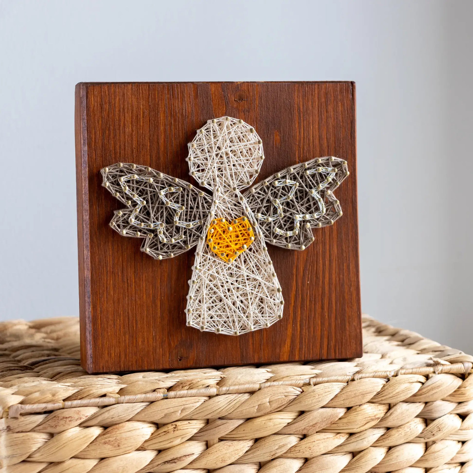 String art angel on a wooden block with a yellow center, placed on a woven surface.