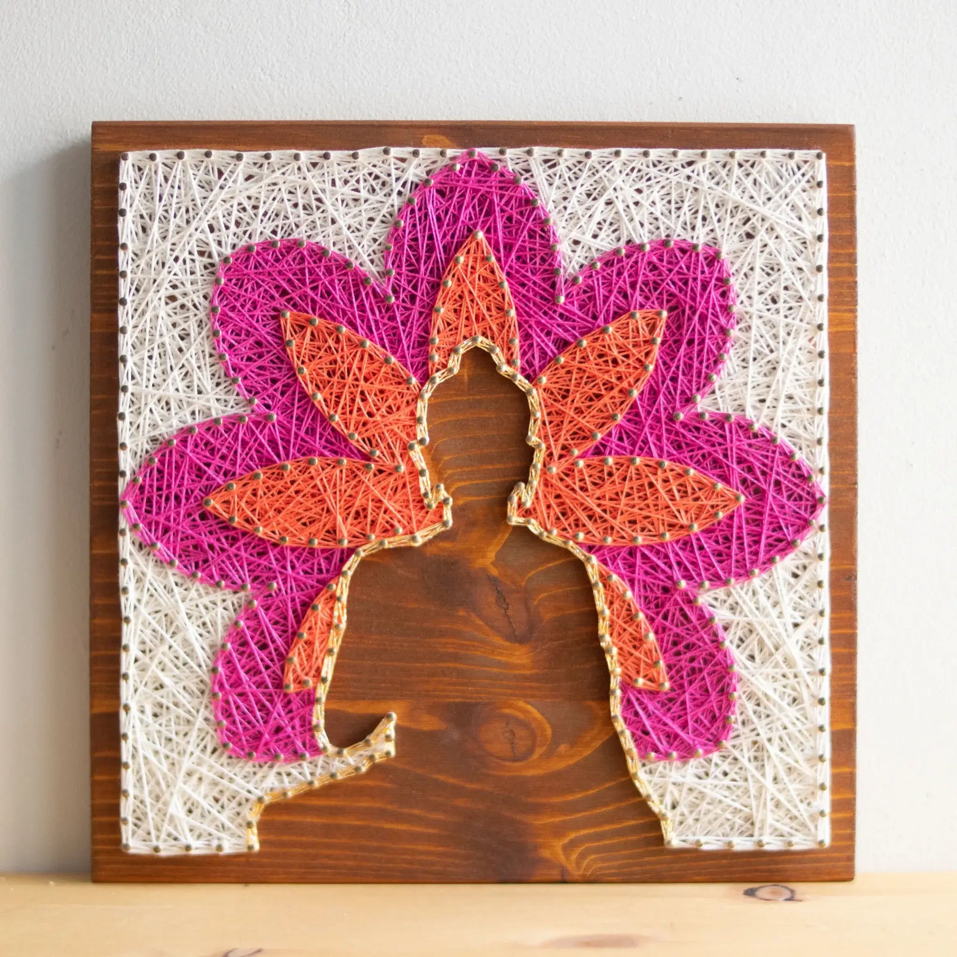 Front view. Buddha silhouette string art with vibrant lotus petals in pink, orange, and white, mounted on a wooden background, representing a peaceful, meditation-inspired design.