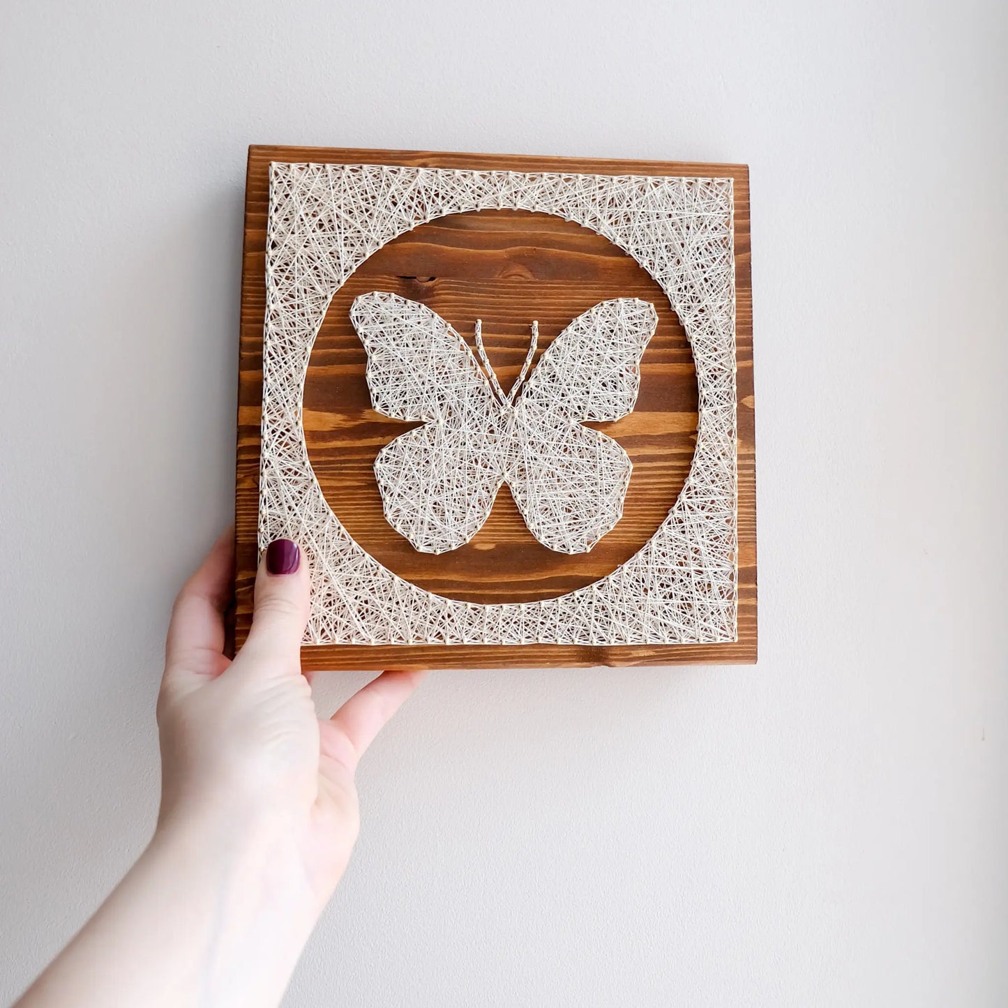 Hand holding butterfly string art, completed project from a craft kit for adults, featuring white string design on a wooden plaque.