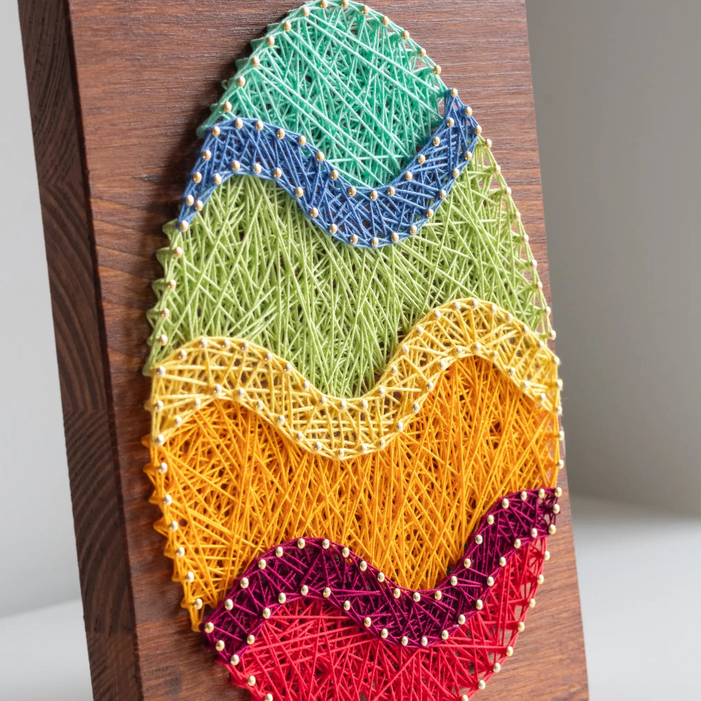 A piece of string art on a wooden board depicting an Easter egg. The egg is created using colorful strings and gold pins, with wavy stripes in different colors: turquoise at the top, followed by blue, green, yellow, orange, purple, and red at the bottom.