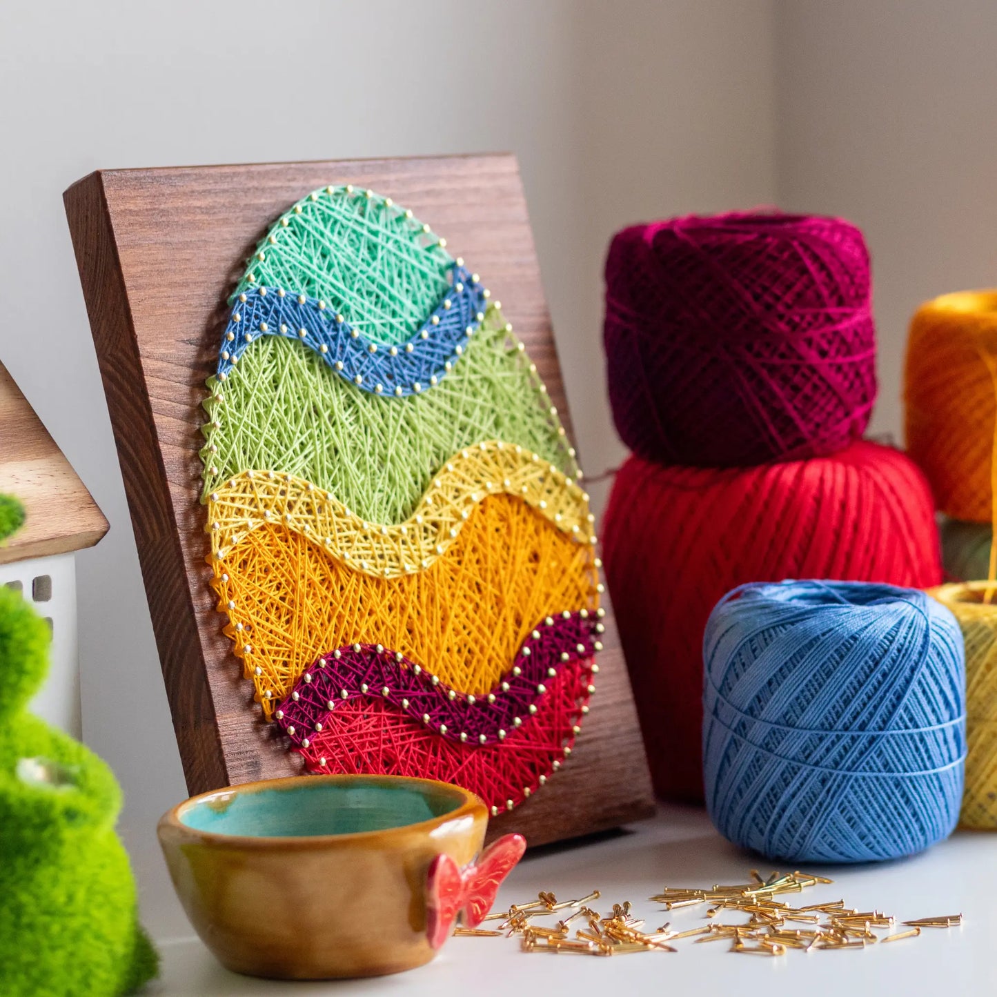 A piece of string art on a wooden board depicting an Easter egg. The egg is created using colorful strings and gold pins, with wavy stripes in different colors: turquoise at the top, followed by blue, green, yellow, orange, purple, and red at the bottom.