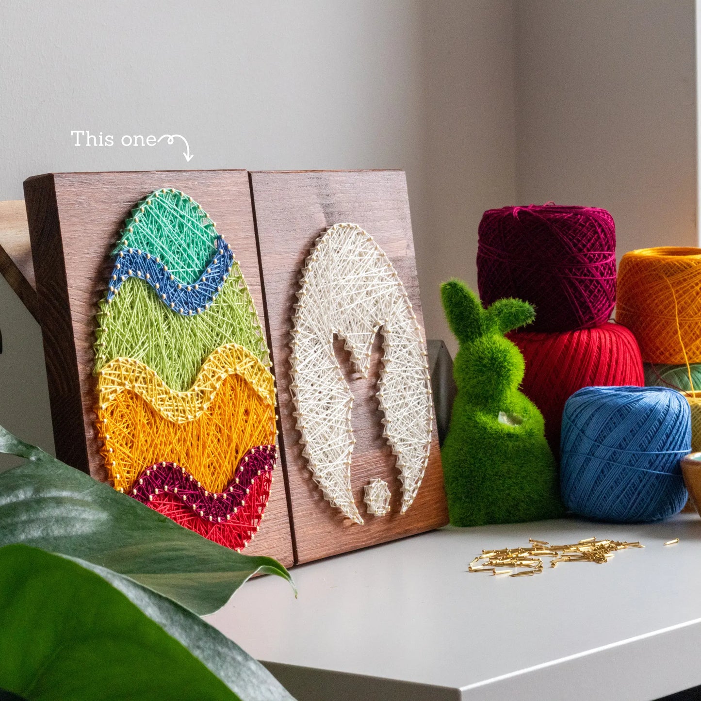 A piece of string art on a wooden board depicting an Easter egg. The egg is created using colorful strings and gold pins, with wavy stripes in different colors: turquoise at the top, followed by blue, green, yellow, orange, purple, and red at the bottom.