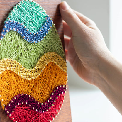 A piece of string art on a wooden board depicting an Easter egg. The egg is created using colorful strings and gold pins, with wavy stripes in different colors: turquoise at the top, followed by blue, green, yellow, orange, purple, and red at the bottom.