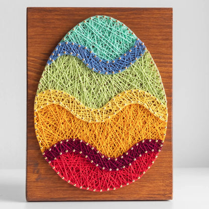 A piece of string art on a wooden board depicting an Easter egg. The egg is created using colorful strings and gold pins, with wavy stripes in different colors: turquoise at the top, followed by blue, green, yellow, orange, purple, and red at the bottom.