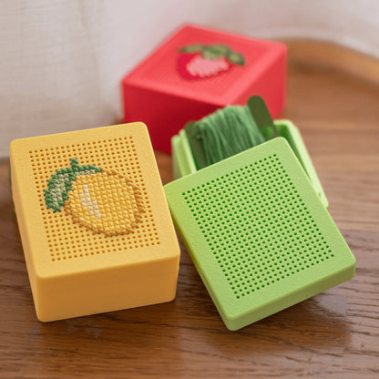 Yellow and green stitchable cross stitch boxes on a wooden surface, one finished with a lemon embroidery and one shown blank, with thread stored inside the box