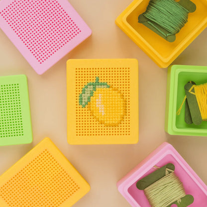 Yellow stitchable cross stitch box lid with a stitched lemon design, surrounded by pink, green, and yellow embroidery boxes and thread spools in a flat lay arrangement