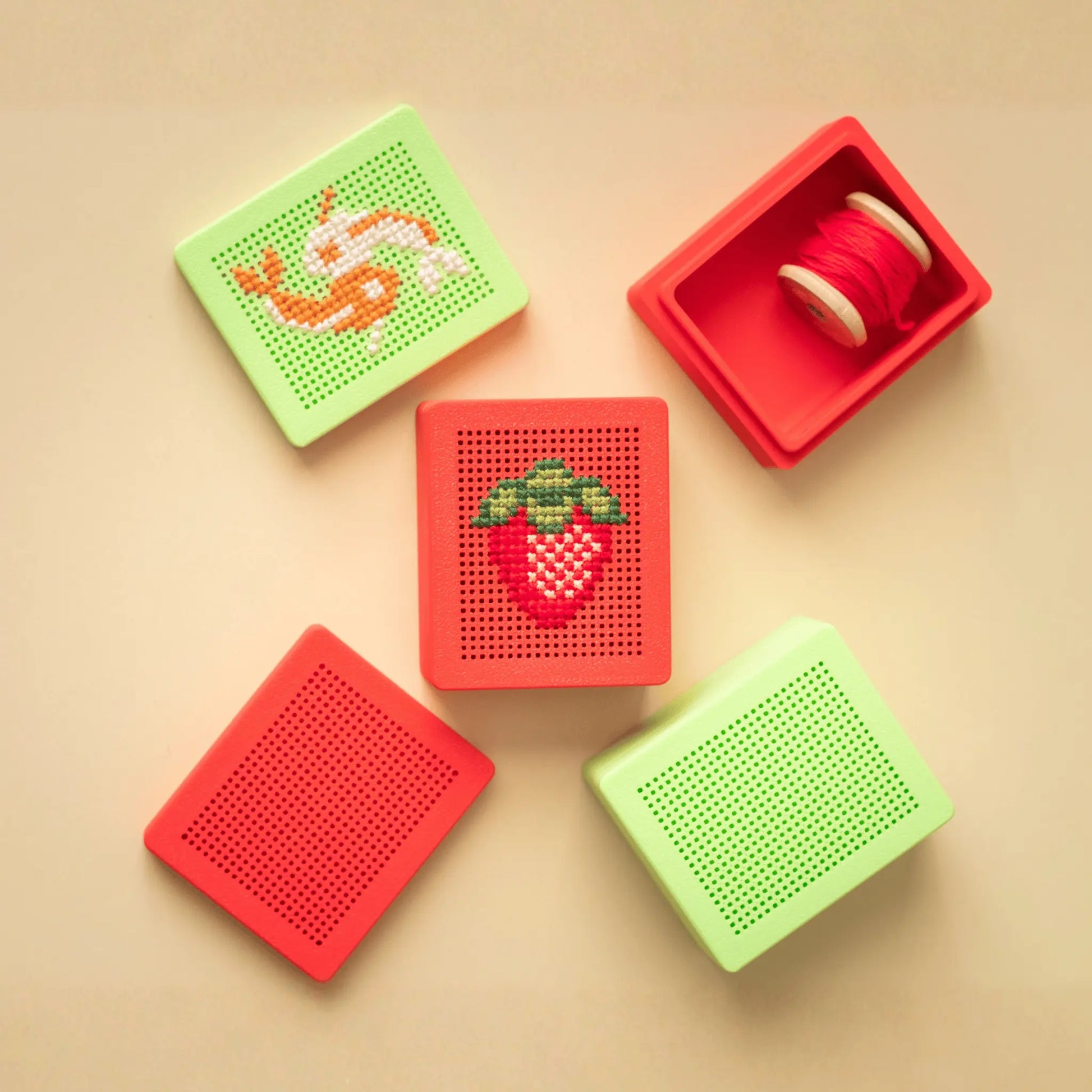 Flat lay of colorful stitchable cross stitch boxes, including red and green embroidery box blanks with stitchable grid lids, one box open with thread inside, and examples of finished cross stitch designs on the lids.