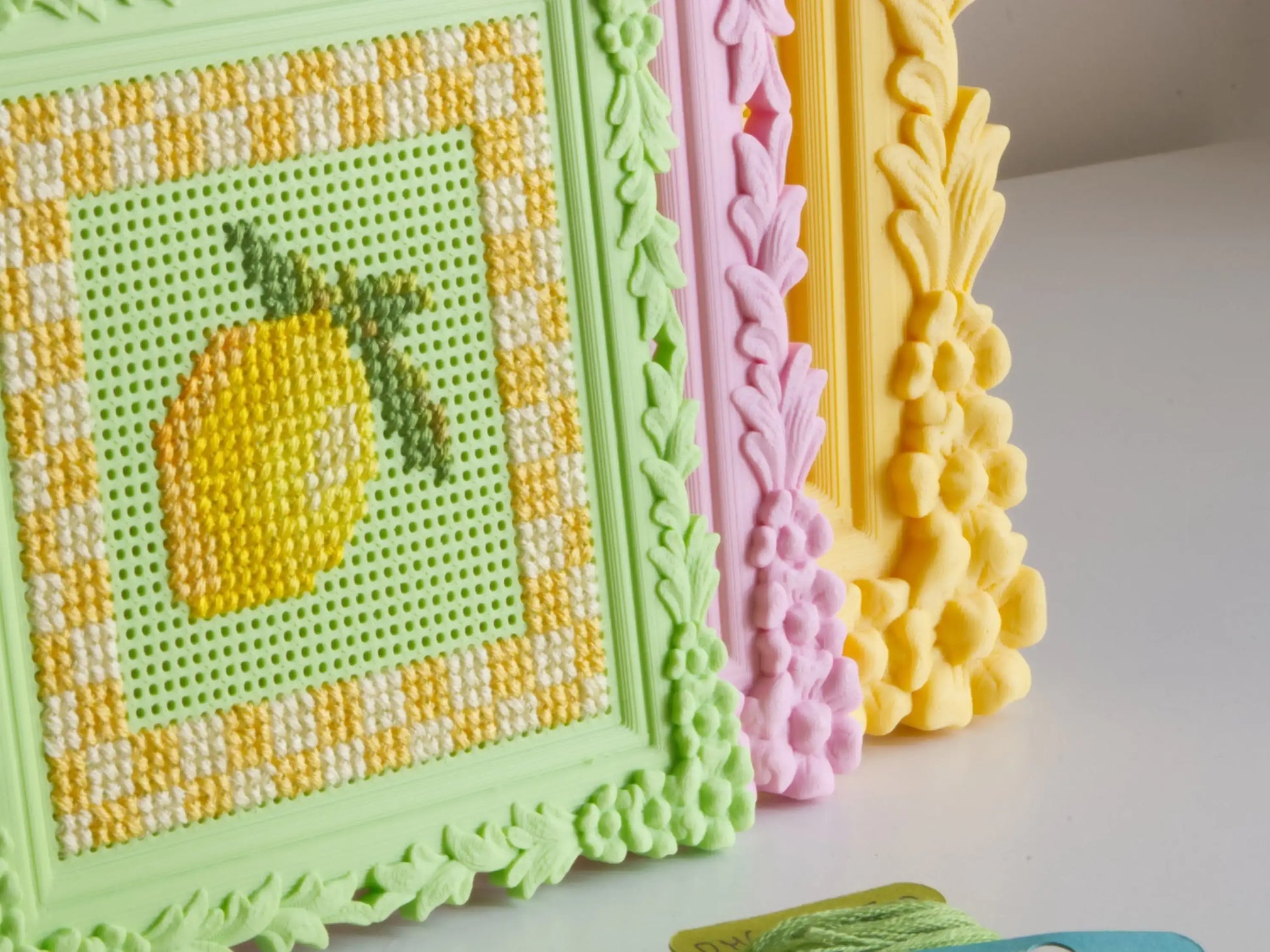 Close-up of a green 3D-printed ornate frame with a perforated stitching grid, embroidered directly with a yellow lemon and gingham border. Pink and yellow floral frames are layered behind it.