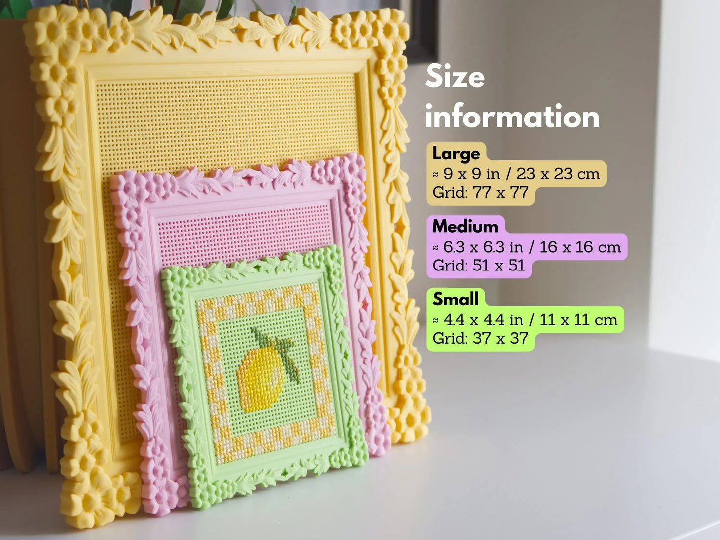 Three ornate square 3D-printed cross stitch frames stacked to compare the large yellow, medium pink, and small green sizes. The small frame features a stitched lemon, while the larger grids are blank.