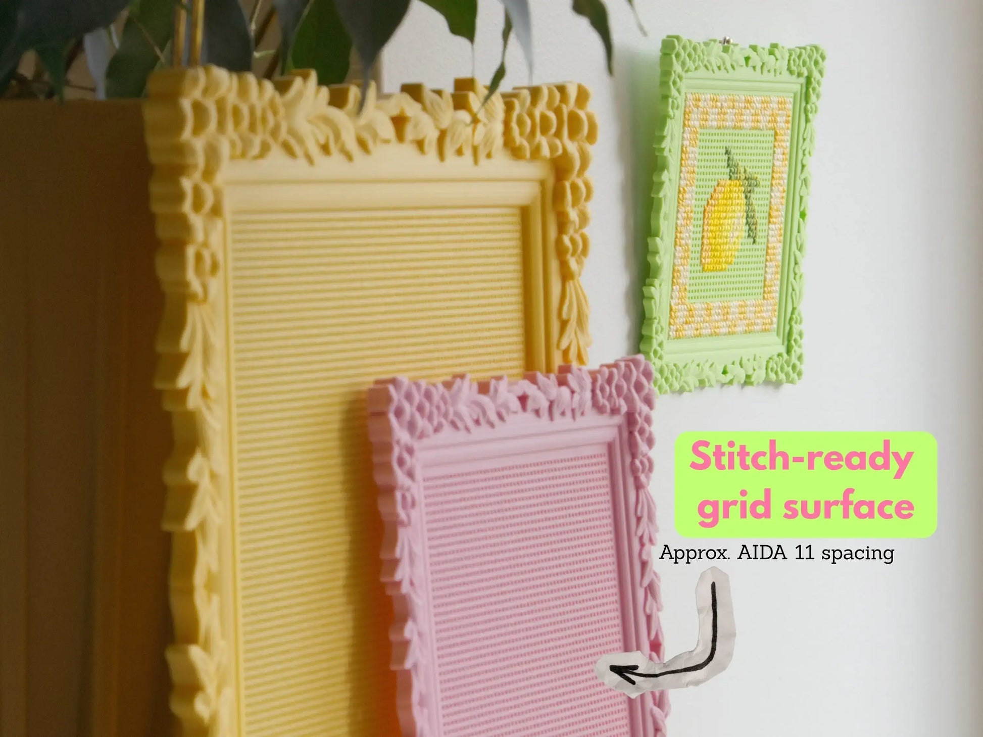 Three ornate 3D-printed cross stitch frames in yellow, pink, and green, shown with built-in perforated grids. The green frame is stitched with a lemon. Text reads “Stitch-ready grid surface” and “Approx. AIDA 11 spacing.”