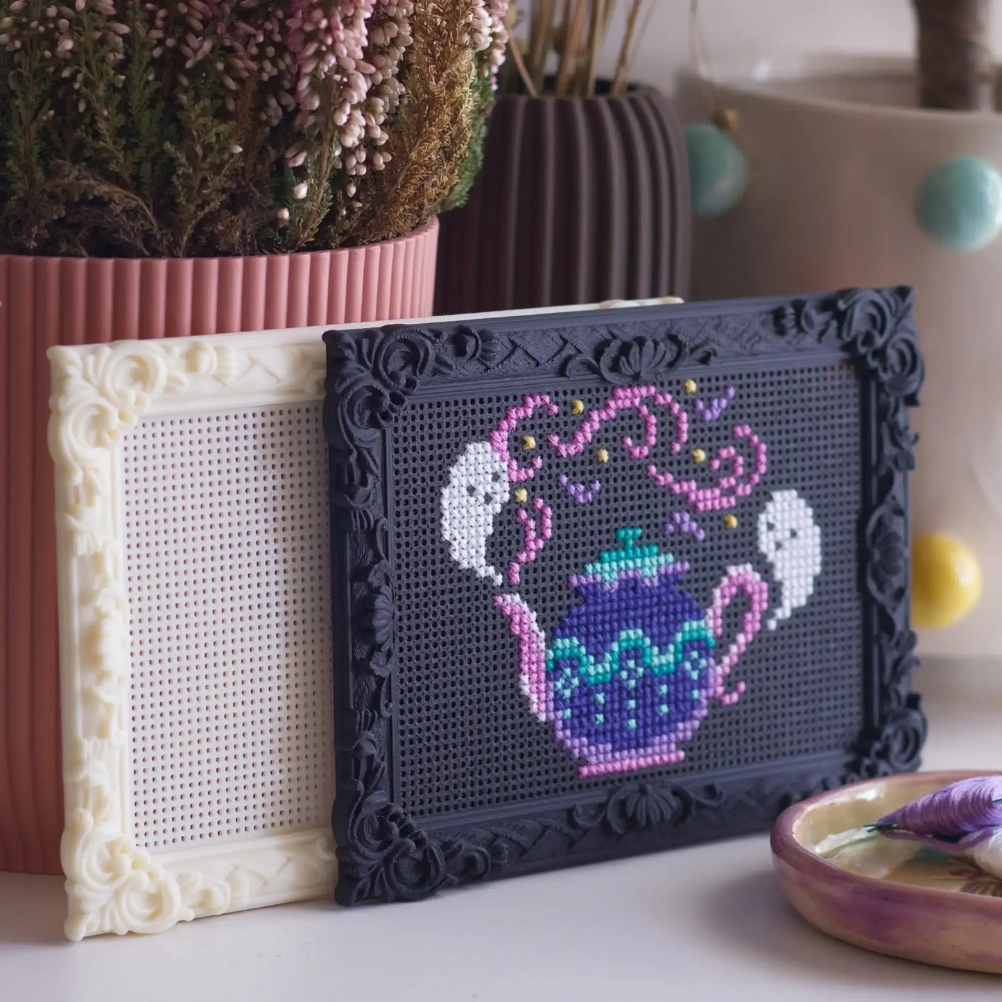 Black baroque rectangle cross stitch frame with a finished ghost teapot design, next to a blank cream frame, styled with dried flowers and a trinket dish.