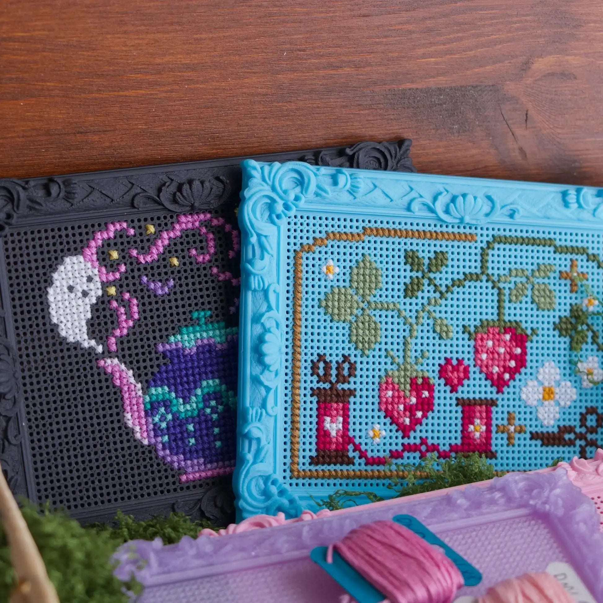Black and blue baroque rectangle cross stitch frames with finished ghost teapot and strawberry designs, shown on a wood surface with pink embroidery floss.