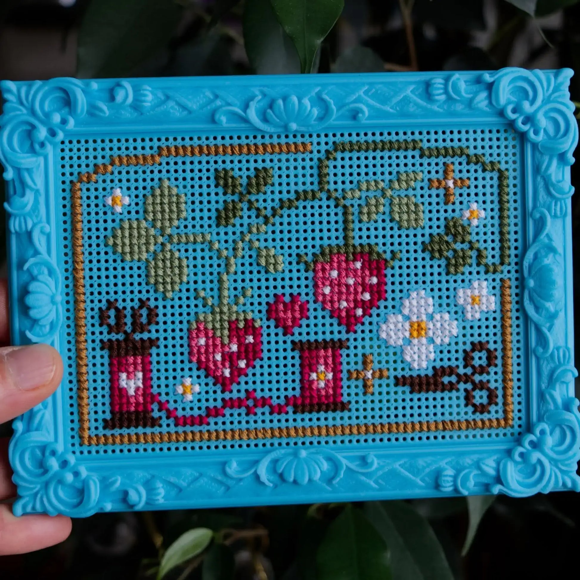 Blue baroque rectangle cross stitch frame held in hand, with a finished strawberry, flower and thread-spool design, against dark green foliage.