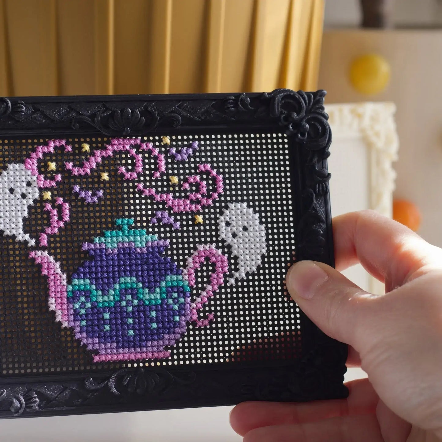 A hand holding a black ornate frame with a cross-stitch style tea-themed design featuring a purple teapot, pink steam, and white ghosts on a dark grid background.