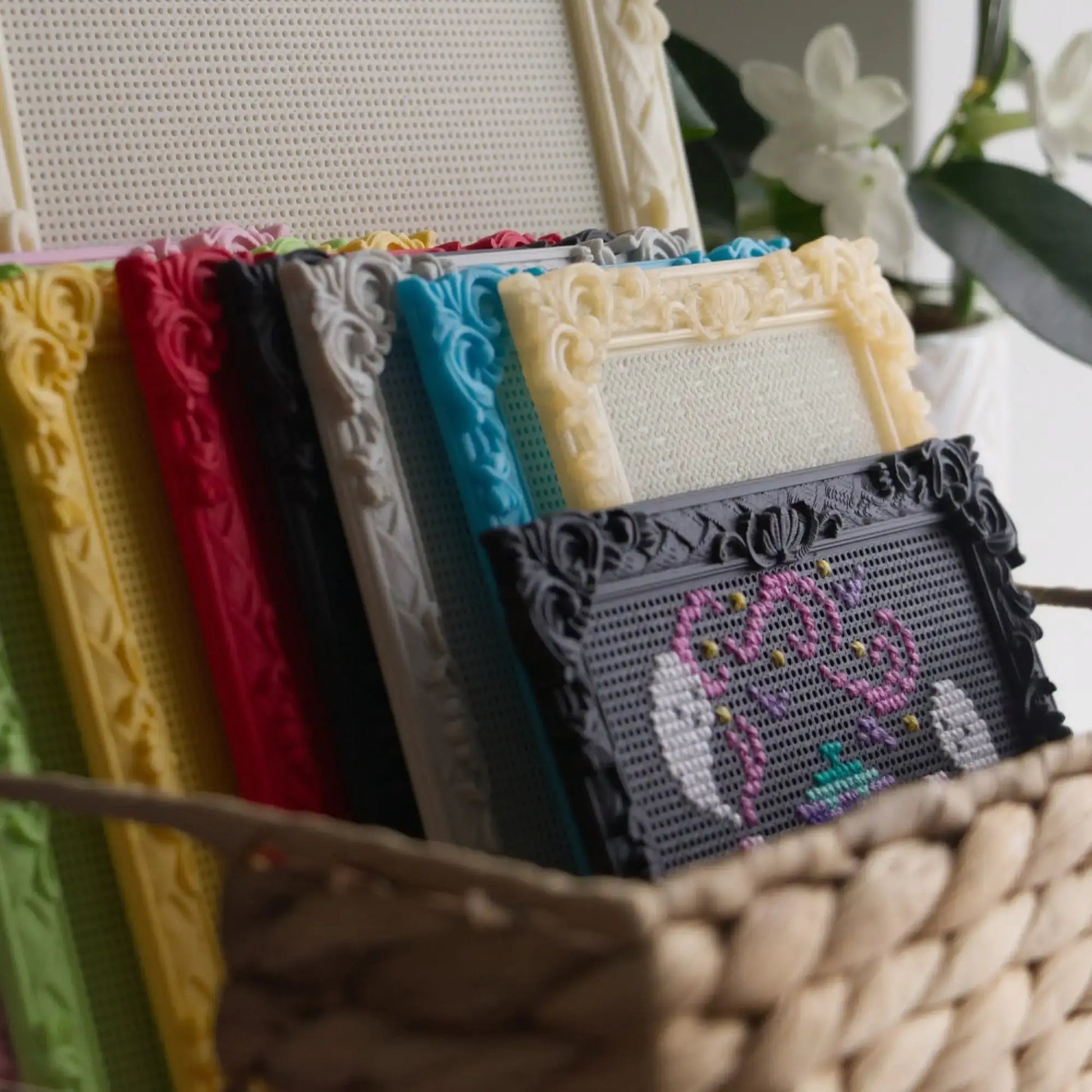 Baroque rectangle cross stitch frames in yellow, red, black, grey, blue and cream stacked in a woven basket, with a finished ghost teapot design in front.