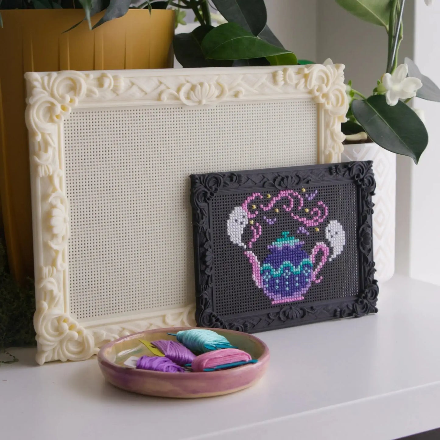 Large blank cream and small black baroque rectangle cross stitch frames, the black one with a finished ghost teapot design, styled with a dish of embroidery floss.