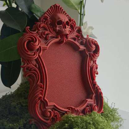 Red marble gothic stitchable cross stitch frame with skull detail, ornate scrollwork, and built-in stitching grid, shown on green moss.