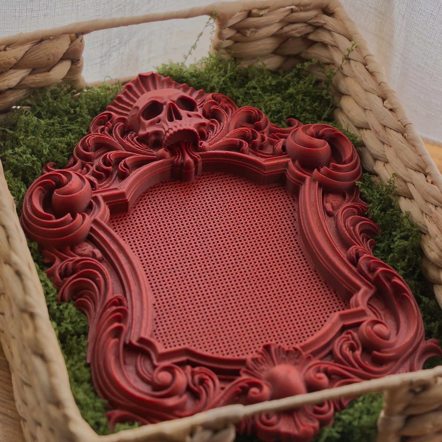 Red marble gothic stitchable cross stitch frame with skull detail, ornate scrollwork, and built-in stitching grid, shown on green moss.