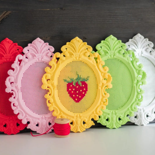 Set of stitchable cross stitch frames in multiple colors, with one finished strawberry cross stitch design and blank frames shown side by side.