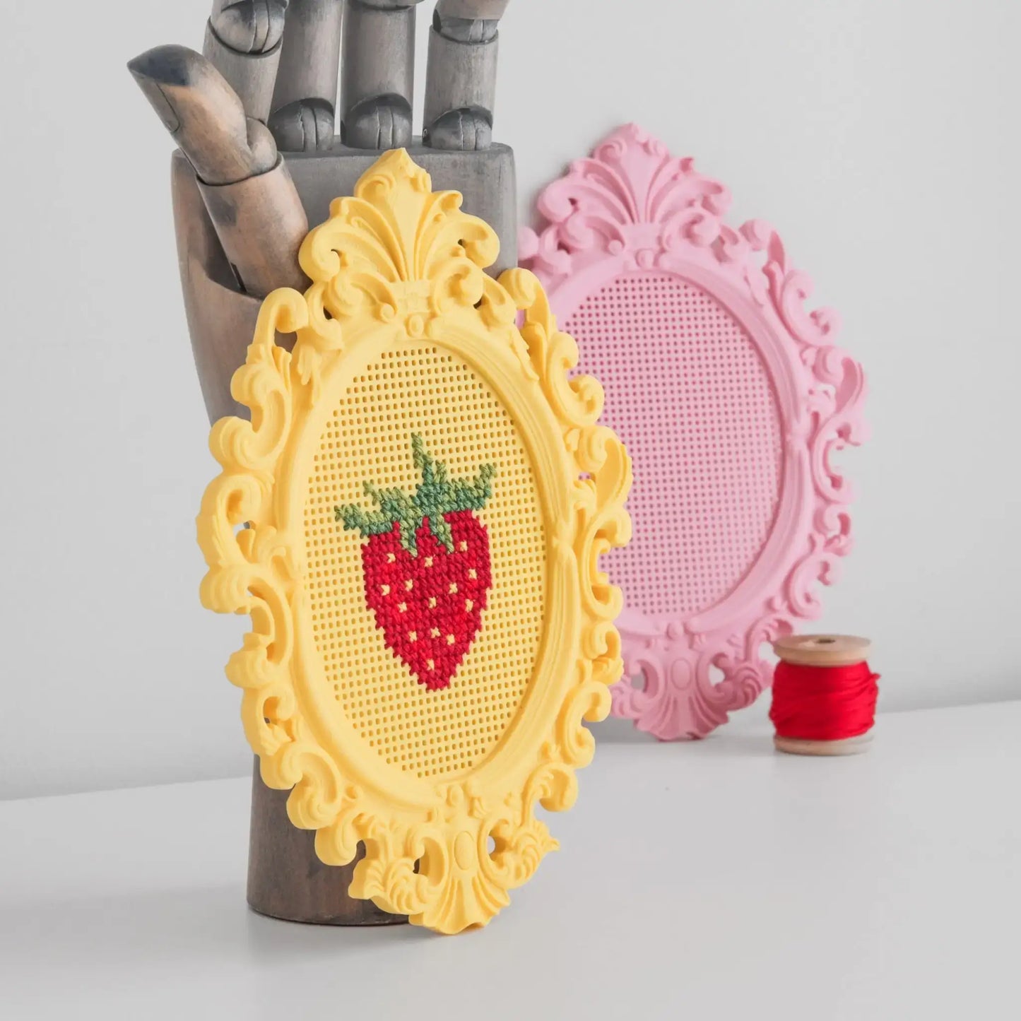 Stitchable cross stitch frame standing upright, showing a finished strawberry cross stitch in a yellow frame with a pink blank frame in the background.