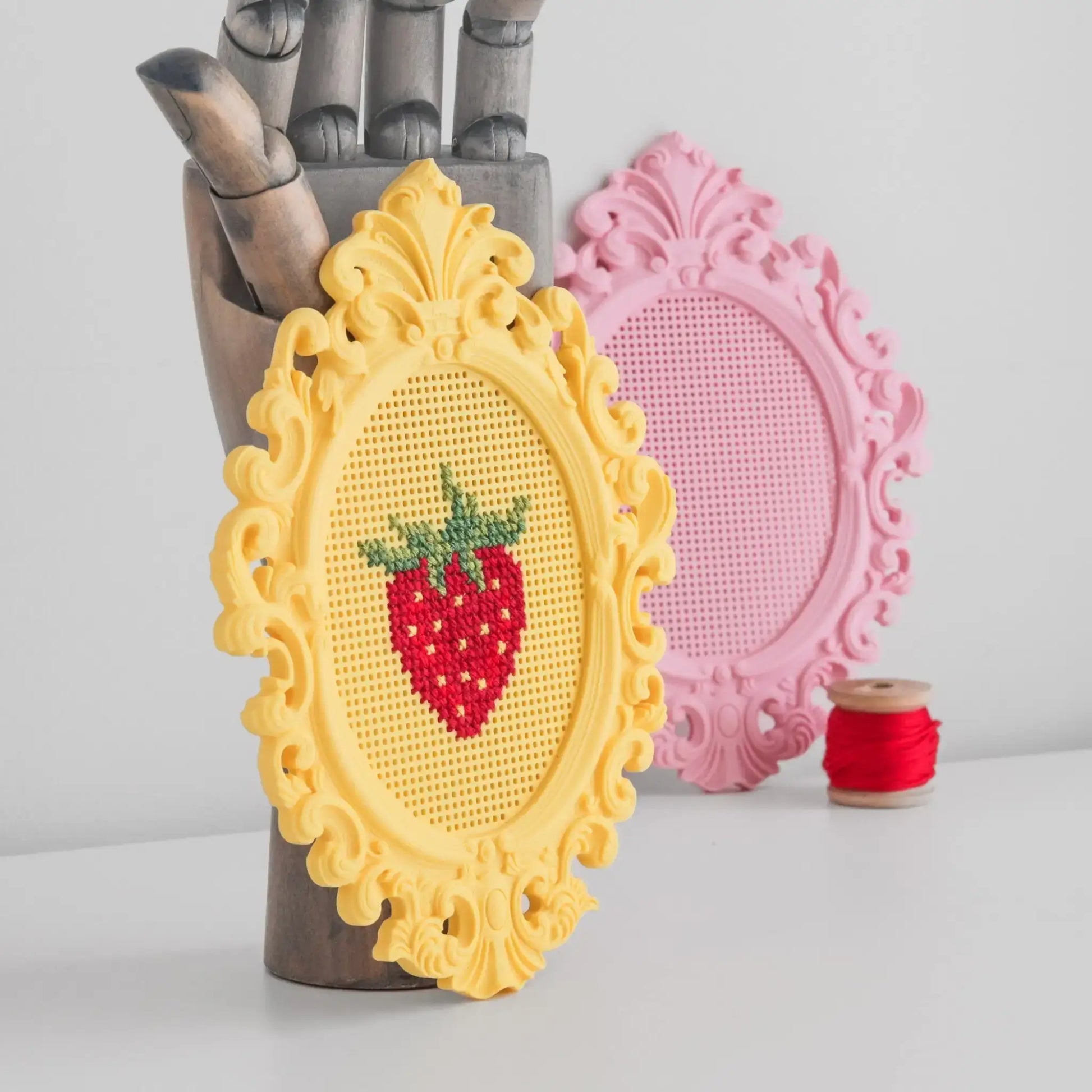 Stitchable cross stitch frame standing upright, showing a finished strawberry cross stitch in a yellow frame with a pink blank frame in the background.