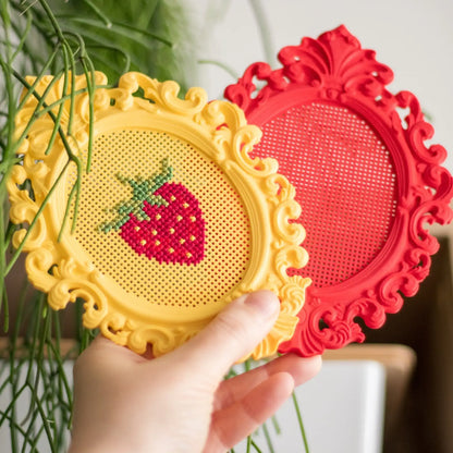 Hand holding two stitchable cross stitch frames, showing a finished strawberry design in a yellow frame and a blank red frame ready for stitching.