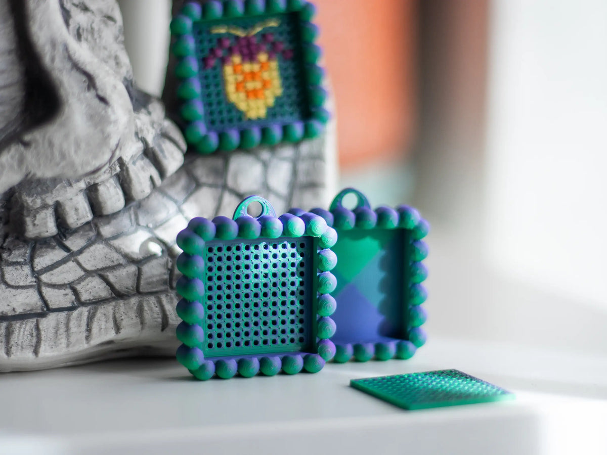 Teal and purple two-tone keychain charms on a bright windowsill: one stitched with a yellow motif, one blank, and an empty base next to its loose grid insert.