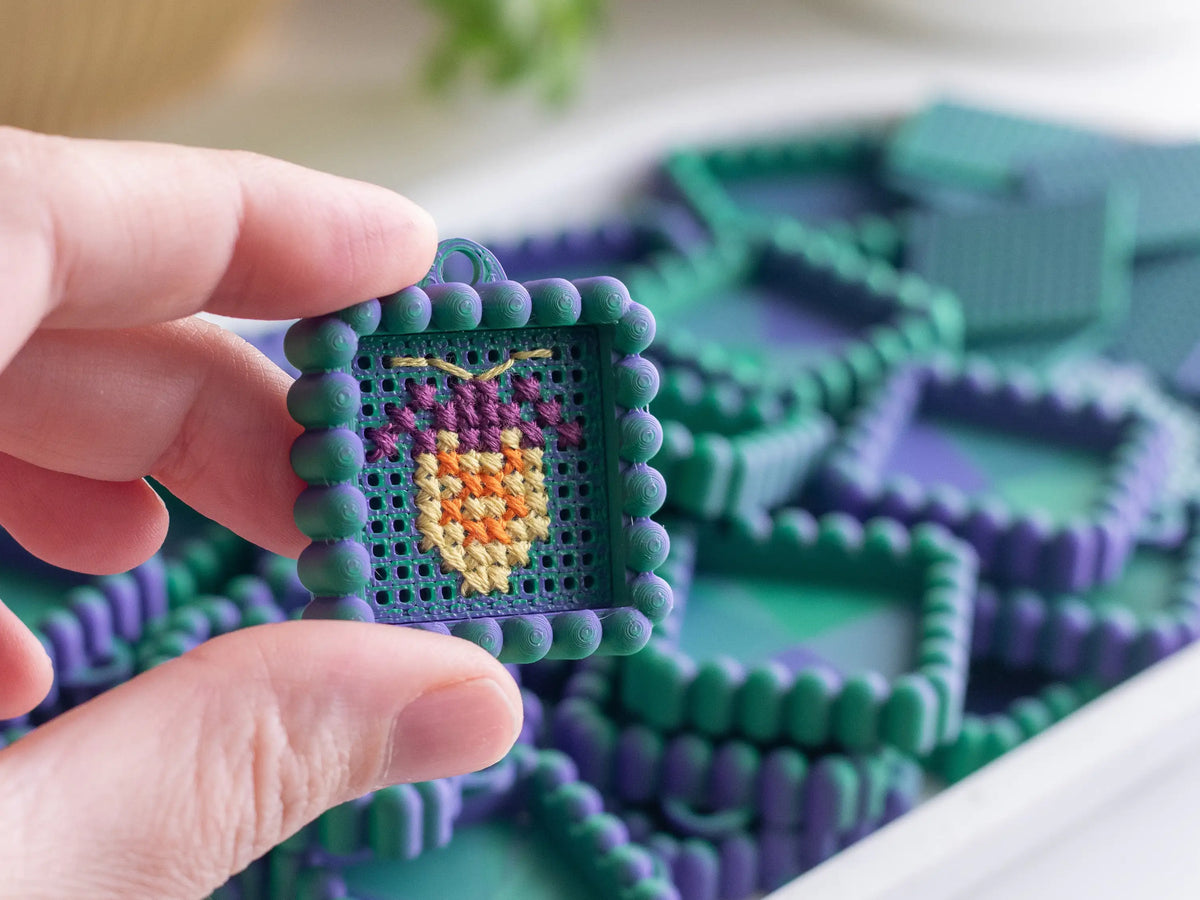 Fingers holding a teal and purple cross stitch keychain with a yellow and orange stitched motif, in front of a tray of more printed blank charms.